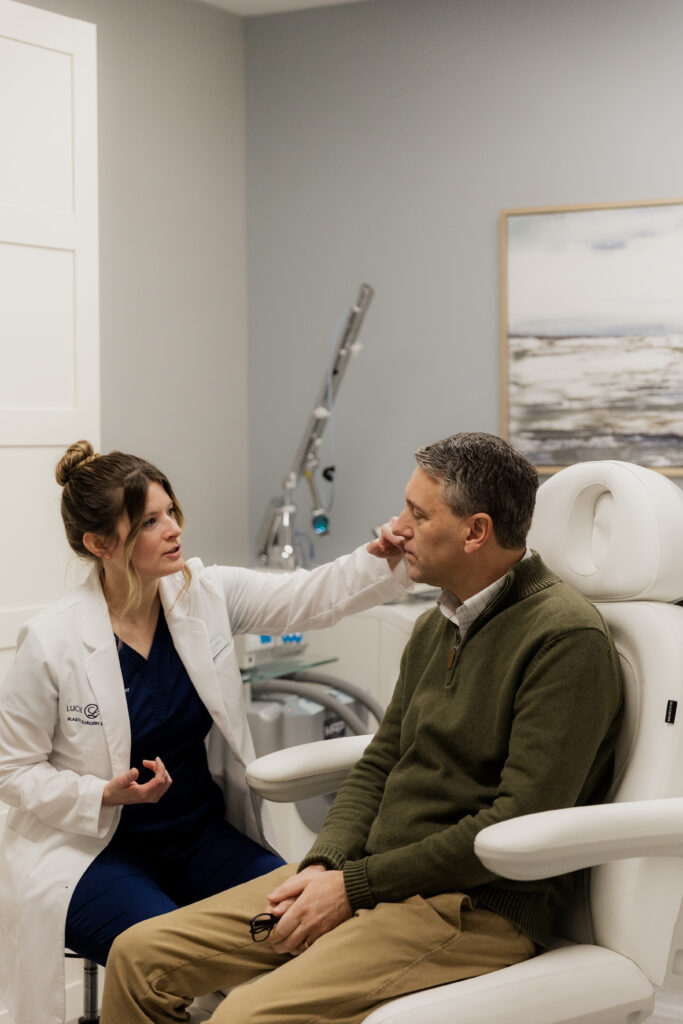 Juliet Coffey, PA-C, performs a consultation in her navy blue scrubs and white coat on a male patient in a dark green sweater. and khakis
